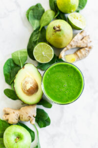 Green smoothie in a glass surrounded by pears, limes, avocados, and other healthy ingredients.